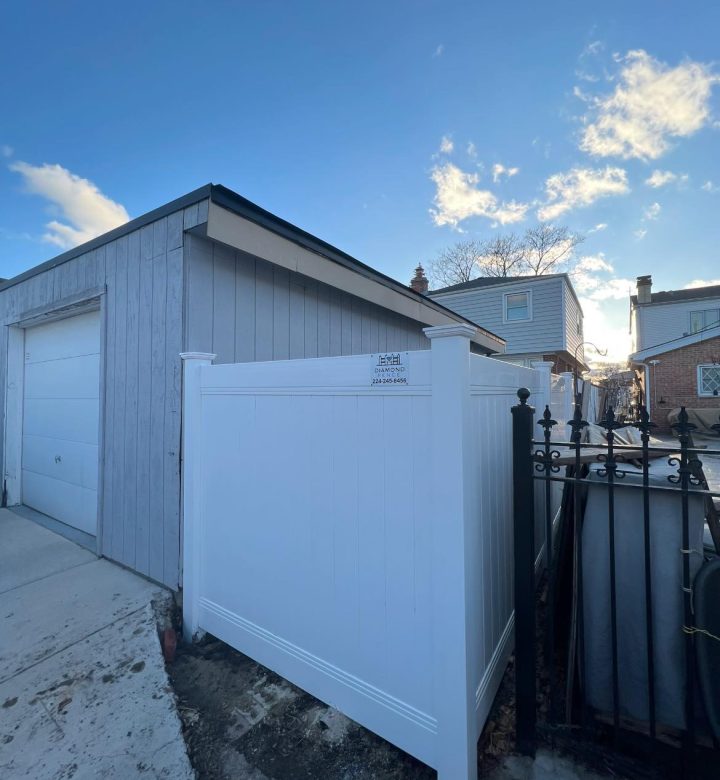 Clean white vinyl privacy fence installed beside gray garage and adjacent to black wrought iron gate — features Diamond Fence sign (224-245-8456) on corner post; perfect for narrow spaces, utility screening, or side-yard privacy under blue sky