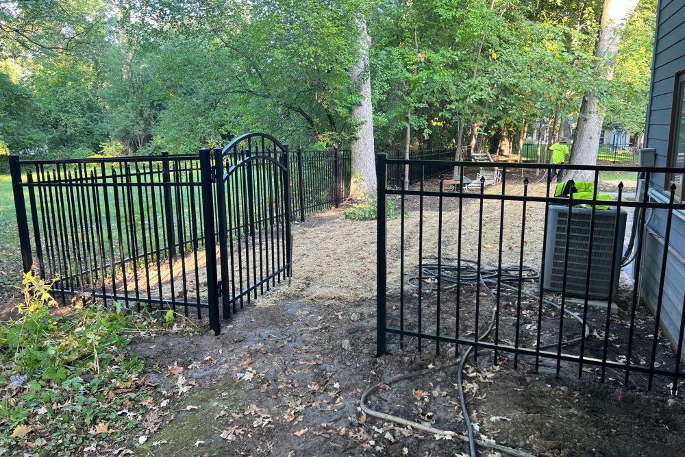Black ornamental aluminum fence with arched gate installed around backyard patio area near AC unit and wooded perimeter
