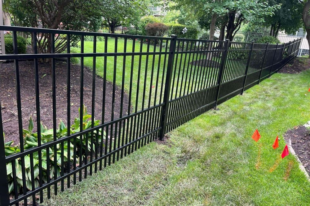 Black aluminum fence following contour of landscaped yard with mulch beds, hostas, and orange utility flags indicating recent installation