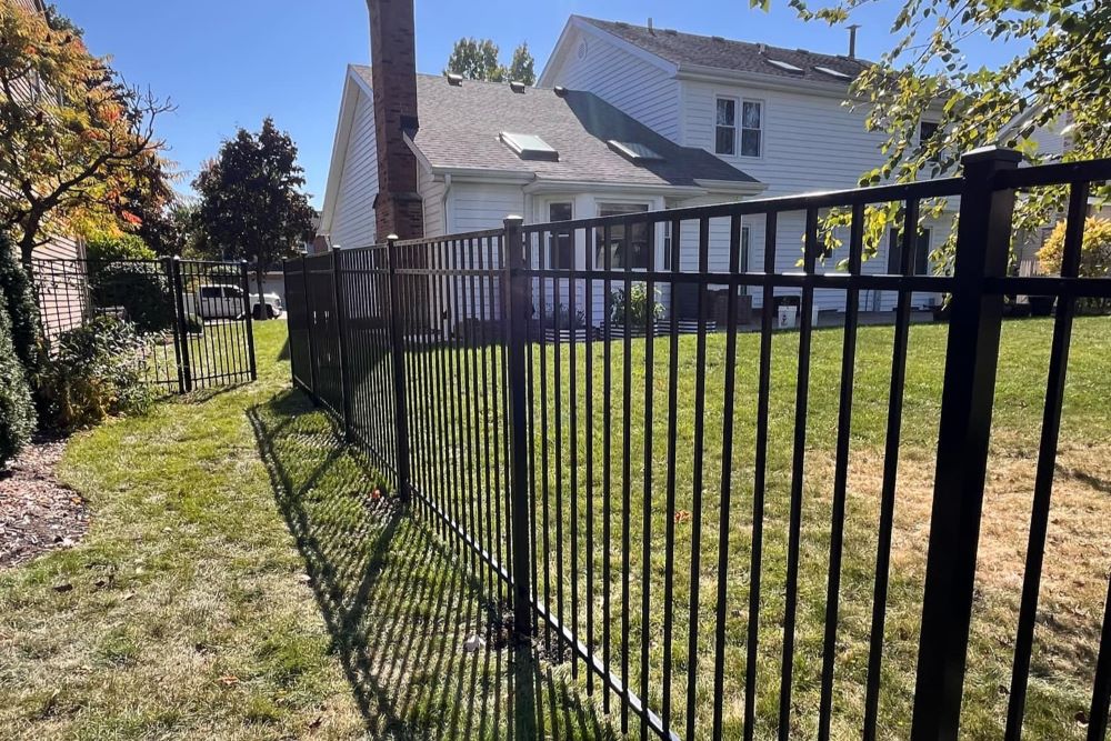 Black aluminum fence enclosing sunny backyard with white house, green lawn, and autumn trees — clean lines and modern curb appeal