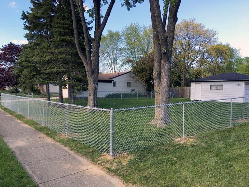 Silver galvanized chain link fence installed along sidewalk with mature trees and homes in background