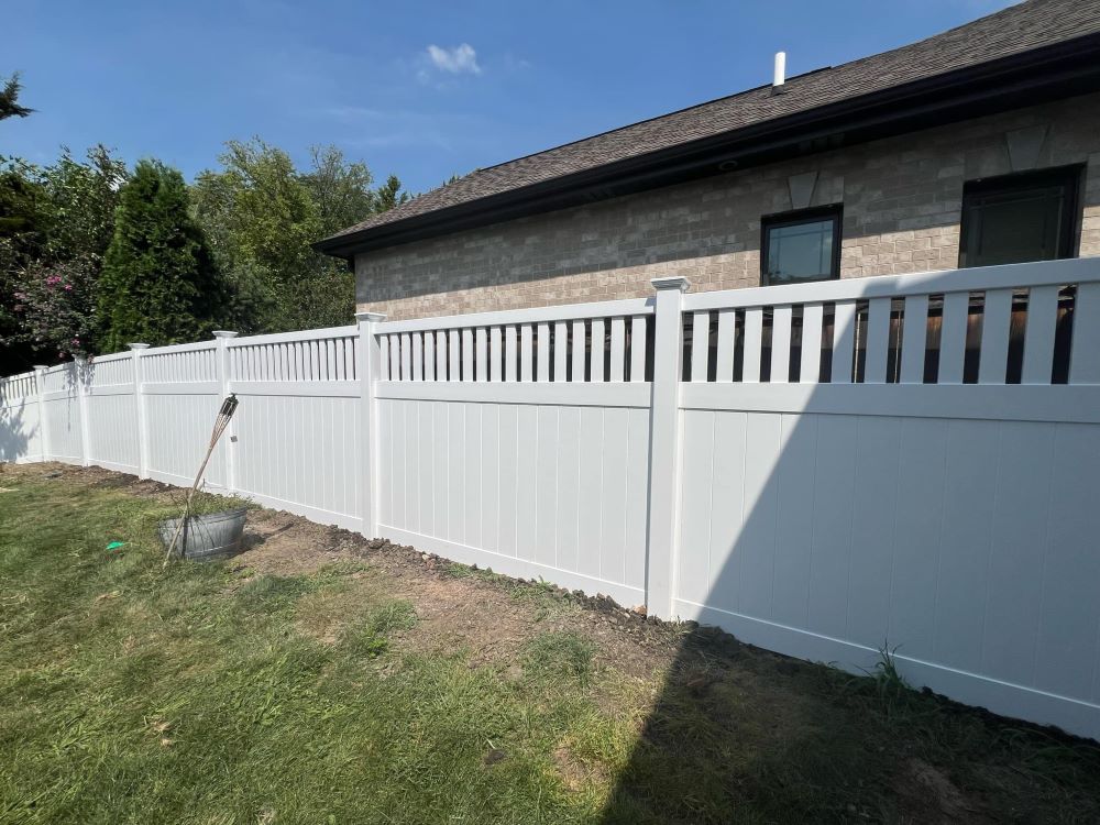Elegant white vinyl privacy fence featuring solid lower panels and vertical picket accent on top section — enhances curb appeal while ensuring full seclusion; shown beside brick home under sunny sky