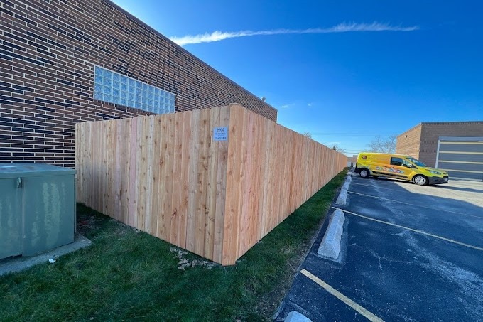 High solid wood privacy fence enclosing commercial building parking lot under clear blue sky — includes utility box concealment and branded signage