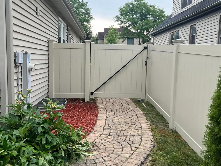 Elegant beige vinyl privacy fence framing curved paver pathway between homes — features black diagonal brace gate, red mulch bed, and lush greenery; perfect for side-yard access and enhanced landscape design