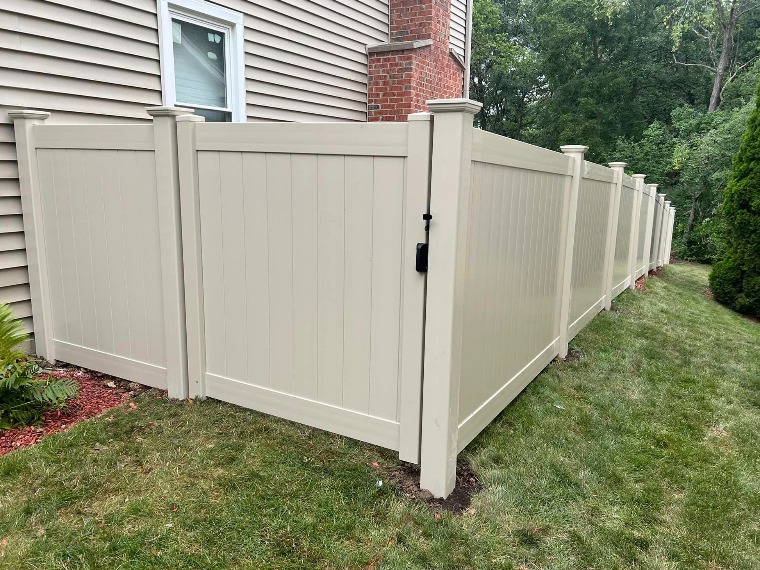 Light beige vinyl privacy fence flanking grassy yard beside house — includes black latch gate, uniform vertical panels, and decorative post caps; ideal for pet containment, noise reduction, and low-maintenance aesthetics