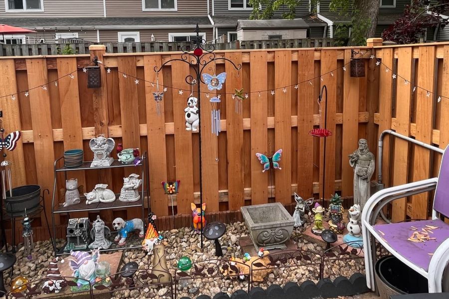 Warm-toned shadowbox wood fence backdrop for whimsical garden display with statues, wind chimes, string lights, and pebble ground cover — perfect for personal outdoor spaces
