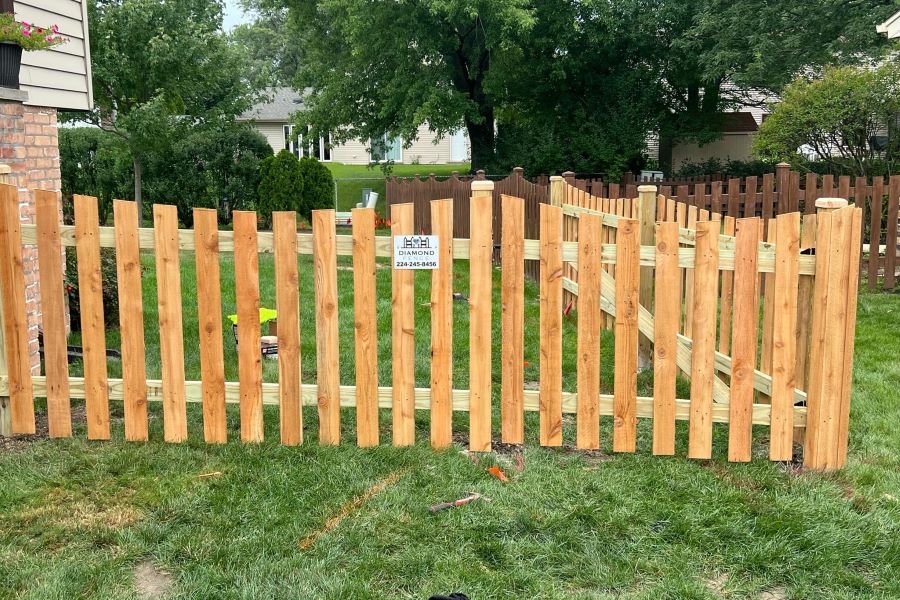 Newly installed natural cedar picket fence with matching gate and horizontal rails, featuring Diamond Fence sign (224-245-8456) on lush green lawn